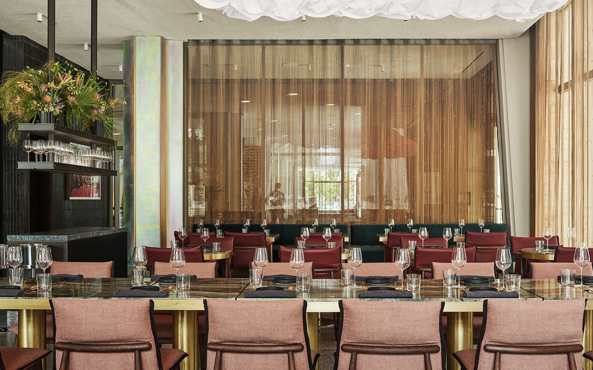 A sophisticated and modern restaurant interior, characterized by a harmony of luxurious textures and colors. The foreground shows tables set with sparkling glassware and black flatware, ready for diners. The tables are flanked by chairs with plush, dusty rose upholstery and golden frames, evoking a sense of refined comfort. A striking feature is the shimmering iridescent panel that vertically accents the space, alongside the soft glow of natural light filtering through sheer, golden-brown curtains. Dark green velvet banquettes line the room, paired with rich, burgundy-colored cushions, creating an atmosphere of opulent warmth. The restaurant's design seamlessly blends contemporary elegance with inviting coziness.
