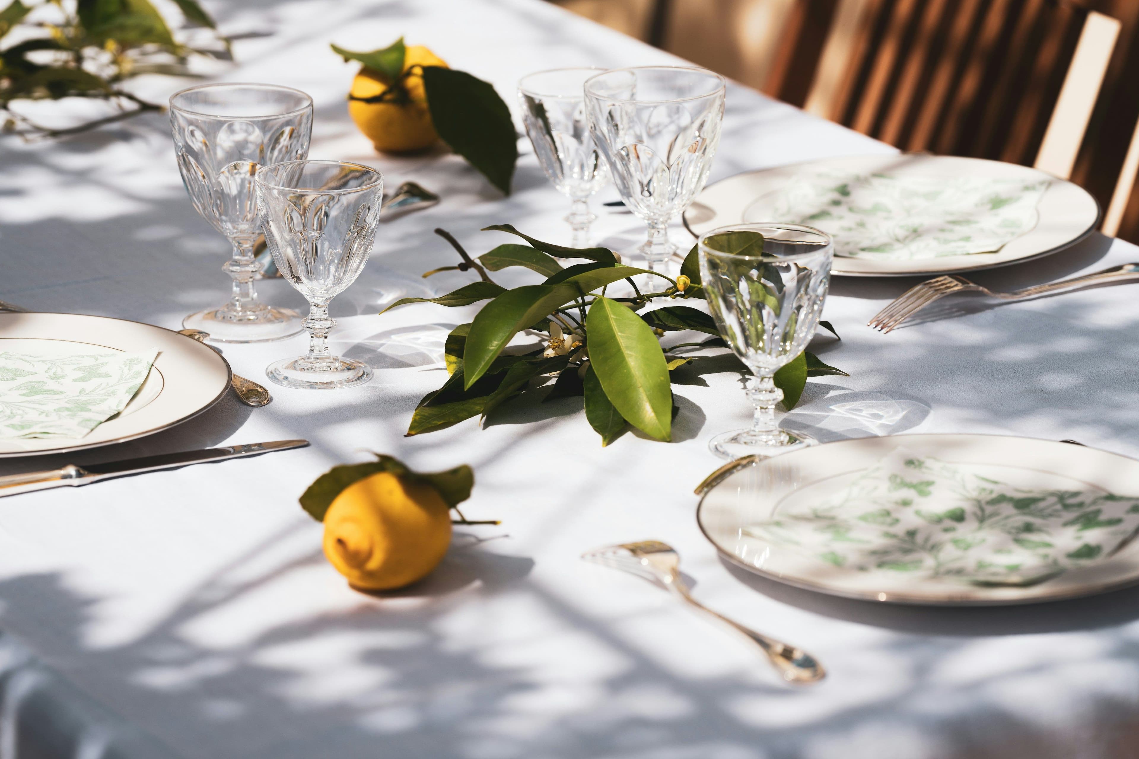 An elegant outdoor dining table setting with crystal glasses, white plates with green leaf patterns, silver cutlery, and fresh lemons on a white tablecloth, representing the fine dining experiences available through Booked in New York.