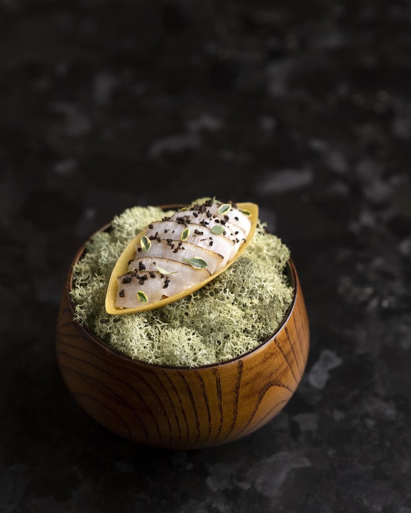Exquisite seafood dish with delicate slices of fish garnished with black sesame seeds and microgreens, elegantly presented on a shell atop a bed of moss in a wooden bowl, embodying the artful cuisine available through New York's top restaurant reservation marketplace.