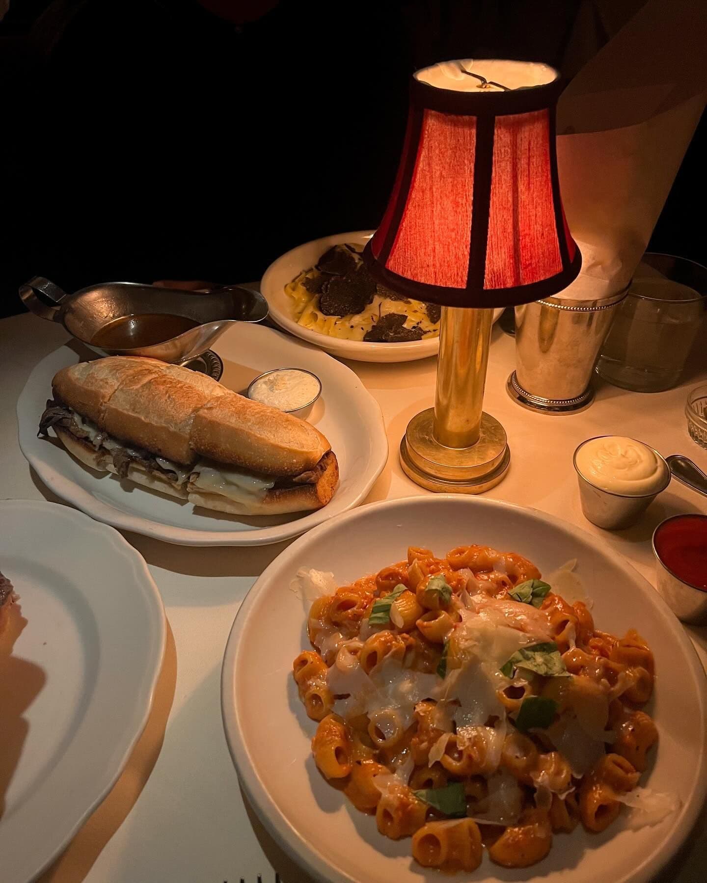An intimate dining setup featuring a steak sandwich and a plate of creamy pasta under the soft glow of a table lamp, offering a cozy and inviting atmosphere as part of the unique dining experiences available through Booked in New York.